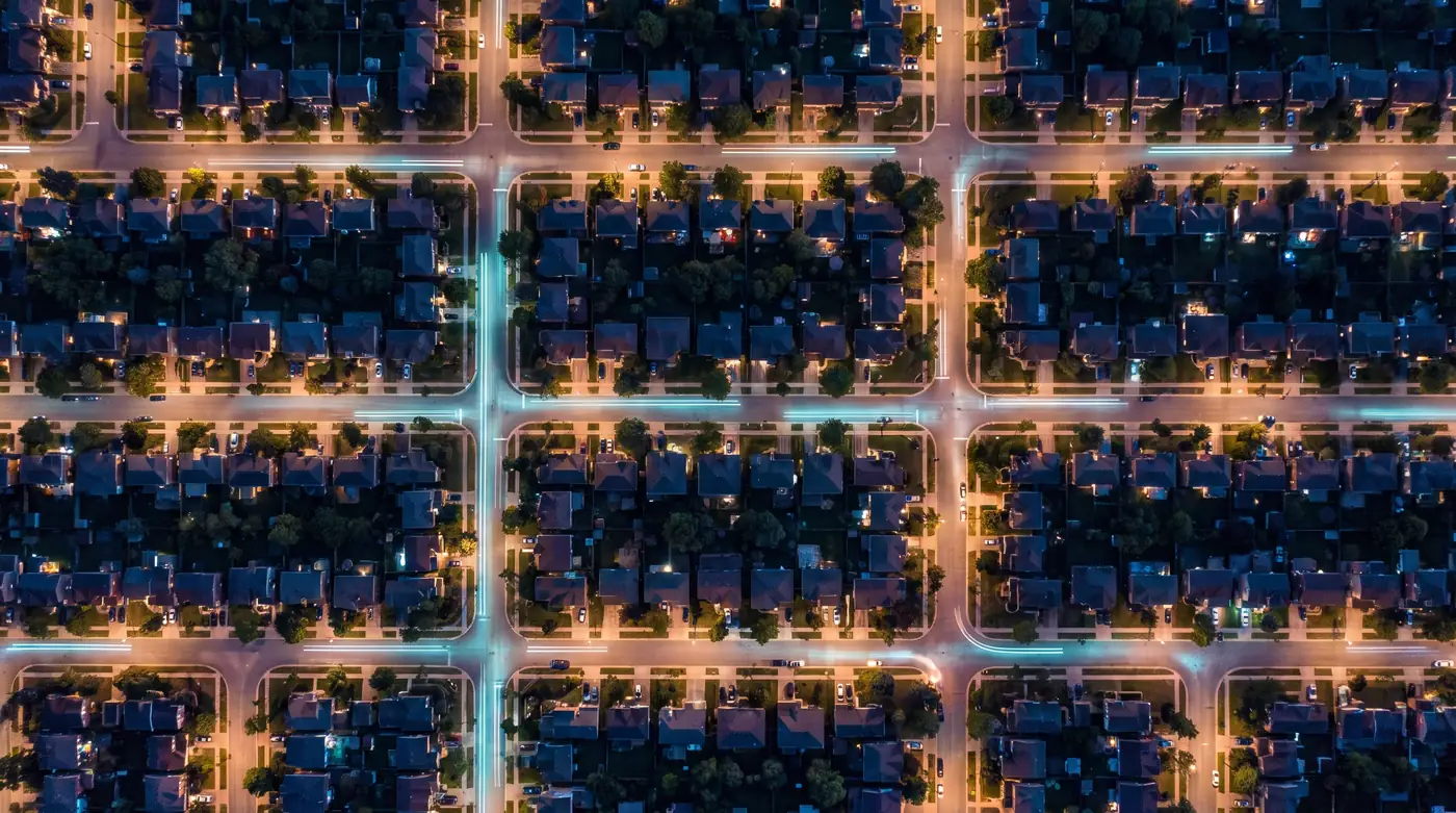 An aerial night view of a suburban neighborhood, warm window lights and street lamps glowing across a grid of streets and rooftops.